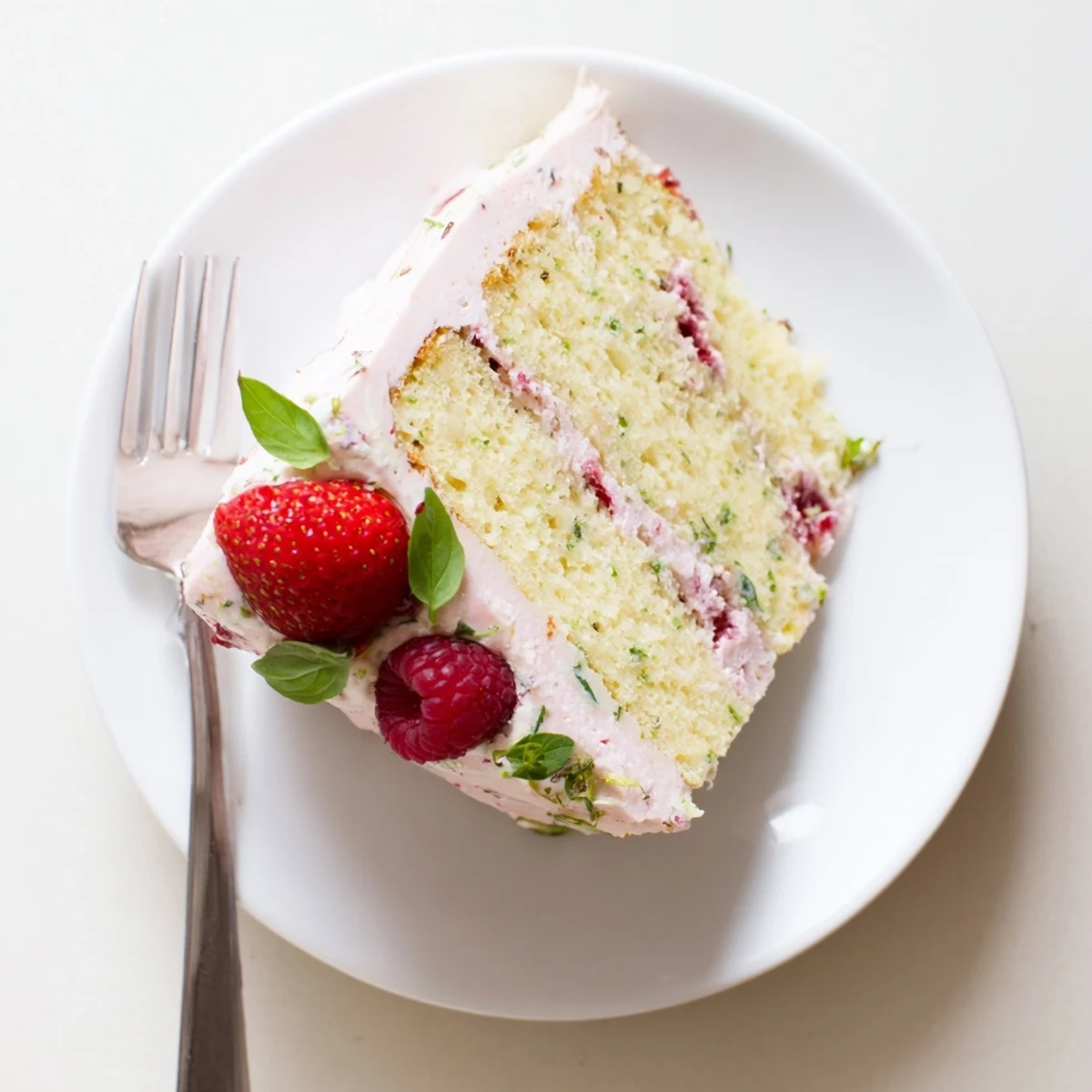 Golden basil berry cake slice topped with creamy basil frosting and fresh mixed berries on white plate